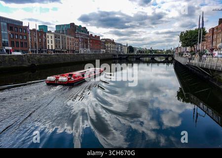 Red Tour Boot fährt auf dem Fluss Liffey in Dublin, Irland, mit Reflektionen von Wolken, historischen Ufergebäuden und Grattan Bridge in der Ferne Stockfoto