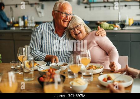 Glücklicher Senior-Mann, der Frau umschließt, während er nach dem Kochkurs am Esstisch zum Mittagessen sitzt Stockfoto