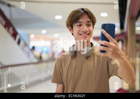 Der 20-jährige Thai-Mann mit Kopfhörern um den Hals schaut auf sein Handy, während er Selfie-Fotos in einem hellen Gang des Einkaufszentrums macht, Nachrichten verschickt und online shoppen kann Stockfoto
