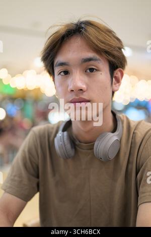 Junger thailändischer Student in einem braunen T-Shirt im Coffee Shop, der Kopfhörer um den Hals trägt, in einem modernen Café-Konzept von sozialen Medien, Fernkommunikation Stockfoto