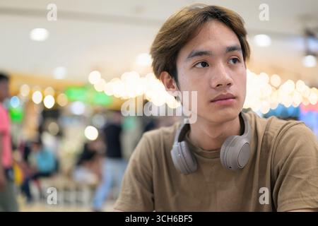 Junger thailändischer Student in einem braunen T-Shirt im Coffee Shop, der Kopfhörer um den Hals trägt, in einem modernen Café-Konzept von sozialen Medien, Fernkommunikation Stockfoto
