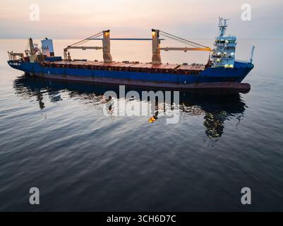 Drohnenansicht eines großen blauen Frachtschiffs mit beleuchteten Decks, das während eines wunderschönen Sonnenuntergangs im ruhigen offenen Meer segelt. Stockfoto
