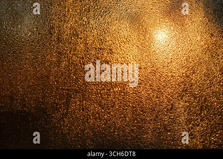 Verschwommene und abstrakte goldene Textur kondensierter Wassertröpfchen auf der Fensteroberfläche, die von hinten durch Sonnenlicht beleuchtet wird. Stockfoto