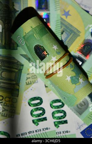 Auf einer Oberfläche verteilte Euro-Banknoten stellen verschiedene Stückelungen der EU-Währung dar. Highlight-Merkmale: Gerollter 100-Euro-Schein, der mit Gummiband gesichert ist und farbenfrohe Spektren und Designelemente zeigt. Stockfoto