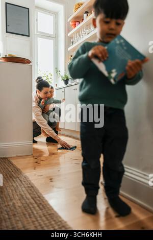 Frau, die den Jungen hält, während sie zu Hause den Boden der Küche reinigt Stockfoto