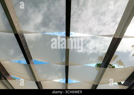 Diagonale Sonnenschirme mit Holzbalken und Blick auf Himmel und Palmenfronten; luftige Pergola mit mediterranem Flair. Stockfoto