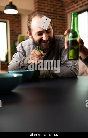 Kaukasier entspannt sich mit Freunden, lächelt mit Karte auf dem Kopf und Bier in der Hand während unterhaltsamer geselliger Zusammenkünfte. Der junge weiße Mann liebt die Bindungszeit und spielt lustiges Ratespiel am Tisch. Stockfoto