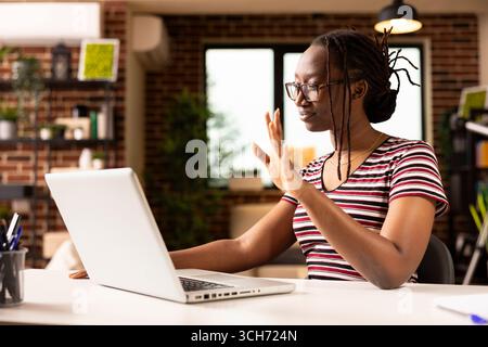 afroamerikanischer Freelancer begrüßt Kunden gerne während eines virtuellen Meetings mit Laptop. Schwarze Geschäftsfrau mit Brille, am Schreibtisch sitzend, während eines Videoanrufs mit einer „Hallo“-Geste an einen Kollegen aus der Ferne. Stockfoto