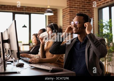 Männlicher afroamerikanischer Mitarbeiter passt sein Headset an, während er sich auf das Gespräch mit Kunden vorbereitet. Ein lächelnder schwarzer Mann hält seine kabelgebundenen Kopfhörer, sitzt am Schreibtisch und kommuniziert mit Kunden aus der Ferne. Stockfoto