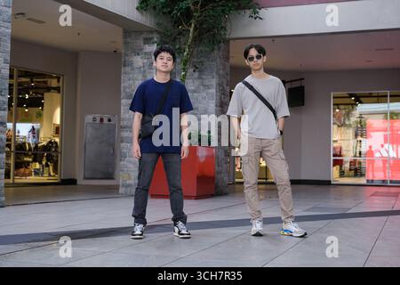 Zwei junge Männer posieren selbstbewusst in einem modernen städtischen Einkaufszentrum. Sie tragen legere Streetwear mit Crossbody-Taschen und Turnschuhen Stockfoto