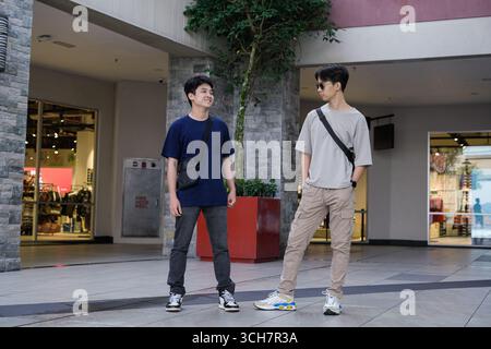 Zwei junge Männer posieren selbstbewusst in einem modernen städtischen Einkaufszentrum. Sie tragen legere Streetwear mit Crossbody-Taschen und Turnschuhen Stockfoto