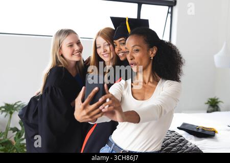 Absolventen in Mützen und Kleidern machen Selfie, feiern gemeinsam im Haus und zu Hause Erfolge Stockfoto