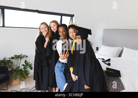 Verschiedene Absolventen tragen Mützen und Kleider, die gemeinsam Selfie im Schlafzimmer machen, feiern, zu Hause Stockfoto