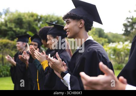 Absolventen verschiedener Studenten in Mützen und Kleidern, die im Garten klatschen und Erfolge feiern Stockfoto