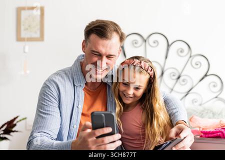 Vater und Tochter lächeln, während sie zu Hause gemeinsam auf das Smartphone schauen Stockfoto