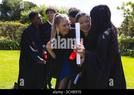 Teenager-Absolventen in Gewändern feiern im Freien, halten Diplome und lächeln fröhlich Stockfoto