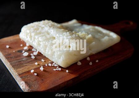 Rohe Lutefisk auf Holzbrett mit grobem Salz, dramatisches Studio-Licht auf schwarzem Hintergrund, minimalistisches Food-Konzept. Stockfoto