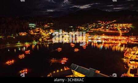 Ein atemberaubender Blick aus der Luft auf ein Dorf am See bei Nacht, beleuchtet mit warmen goldenen Lichtern. Schwimmende Laternen und Boote erzeugen ein faszinierendes Muster o Stockfoto