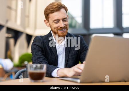 Lächelnder rothaariger Geschäftsmann im Anzug, der im Café im Freien am Laptop arbeitet Stockfoto