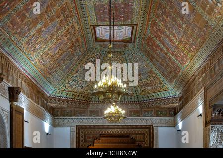 Reich verzierter Empfangsraum mit gemalter Zedernholzdecke, Kronleuchter und gekacheltem Kamin im Bahia Palace (Palais Bahia), Marrakesch, Marokko. Stockfoto
