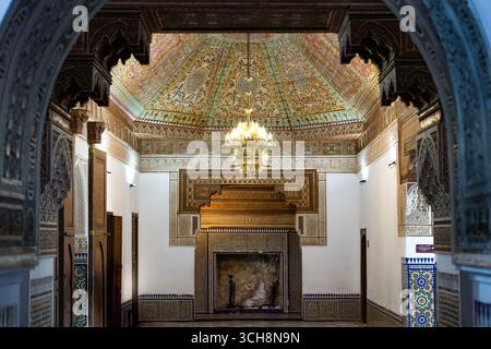 Reich verzierter Empfangsraum mit gemalter Zedernholzdecke, Kronleuchter und gekacheltem Kamin im Bahia Palace (Palais Bahia), Marrakesch, Marokko. Stockfoto