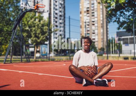Junge afroamerikanische Frau, die auf dem sonnigen städtischen Basketballfeld sitzt und einen Ball mit fröhlichem Lächeln hält, repräsentiert Freude und aktiven Lebensstil. Stockfoto