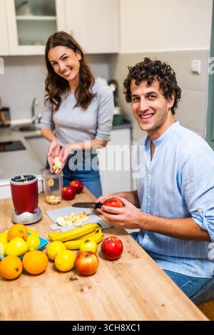 Ein erwachsenes Paar macht Spaß, während es Smoothie in der Küche zubereitet. Sie schneiden Obst und verwenden einen elektrischen Mixer. Stockfoto