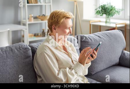 Eine ältere Frau sitzt auf dem Sofa im Wohnzimmer zu Hause und hält das Handy in der Hand. Stockfoto