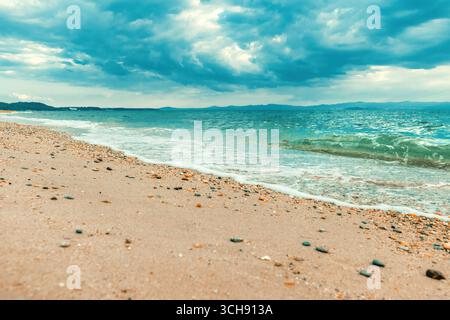 Dramatische Meereslandschaft von Polychrono, Chalkidiki, Griechenland, mit türkisfarbenen Wellen, die unter einem stimmungsvollen Himmel auf einem Kieselstrand schwimmen. Ferne Berge Stockfoto