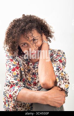 Porträt einer fröhlichen lateinischen Frau mittleren Alters mit lockigen Haaren, die eine bunte Bluse mit Blumenmuster trägt und in einer natürlichen und entspannten Pose sitzt. Studio-Aufnahme Stockfoto