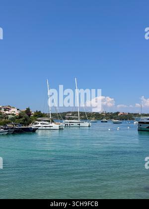 Segelboote legten in einem ruhigen Küstenhafen unter einem hellblauen Himmel an. Klares türkisfarbenes Wasser trifft sanft auf die Boote, mit bezaubernden Häusern Stockfoto