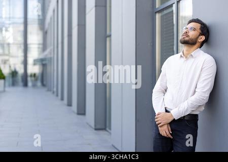 Ein junger Geschäftsmann mit Brille und Bart lehnt sich vor einem Bürogebäude an eine Wand, blickt nachdenklich nach oben und denkt über geschäftliche Angelegenheiten nach Stockfoto