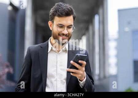 Junger Geschäftsmann mit Brille und Anzug, stehend in einem Stadtgebiet, während er ein Smartphone benutzt, lächelt und auf positive und selbstbewusste Weise mit dem Bildschirm des Geräts interagiert Stockfoto