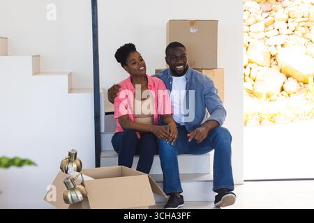 Afroamerikanisches Paar sitzt auf Treppen, packt Kisten aus und lässt sich glücklich in ein neues Zuhause einziehen. Umzug, Umzug, Glück, Lebensstil Stockfoto