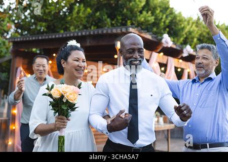 Bei der Hochzeit feiern Sie draußen mit Freunden, ein Seniorenpaar hält Blumen und lächelt fröhlich. Feier, Freundschaft, Glück, Senioren, Bouque Stockfoto