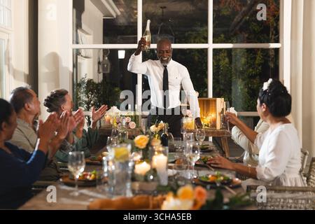 Bei der Hochzeit hob ein älterer Mann beim Familienessen Glas und alle klatschten und lächelten. Feiern, Treffen, Glück, Essen, Jubiläum Stockfoto