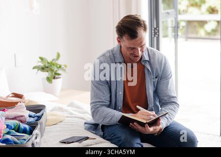 Mann, der in Notizbuch schreibt, während er zu Hause Koffer packt und sich auf die Reise vorbereitet. Reise, Urlaub, Gepäck, Planung, Vorbereitung, Reise Stockfoto
