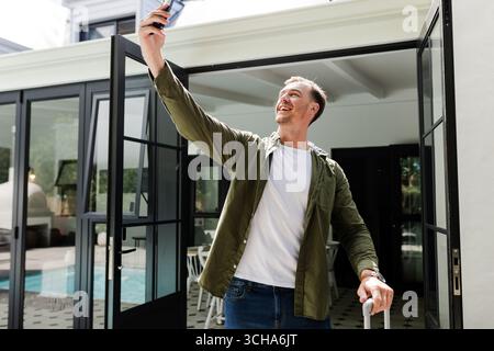 Selfie mit Smartphone machen, Mann steht vor einem modernen Zuhause und lächelt glücklich. Technologie, Architektur, Glück, Lifestyle, Outdoor, Hochformat Stockfoto