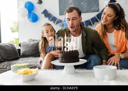 Vater bläst Kerzen auf Geburtstagskuchen aus, während die Familie zu Hause feiert. Feier, Party, Glück, Erziehung, Zweisamkeit, Freude Stockfoto