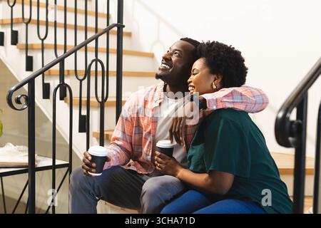 Sitzendes afroamerikanisches Paar trinkt Kaffee auf Holztreppe drinnen und hält Tassen zum Mitnehmen. Gemütlich, Gesellschaft, Lebensstil, warm, Freundschaft, Stockfoto