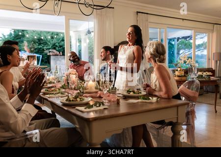 Die Braut hält eine Rede am eleganten Esstisch, die Gäste applaudieren und lächeln herzlich. Hochzeit, Feier, Empfang, Toasten, herzlichen Glückwunsch, Sammeln Stockfoto
