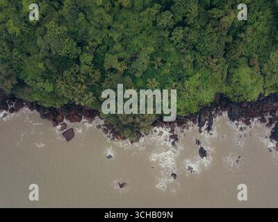 Aus der Vogelperspektive auf die zerklüftete Küste, wo der smaragdgrüne Dschungel auf den matschigen Ozean trifft, die Wellen gegen die dunklen Felsen krachen, Canacona, Goa, Indien. Stockfoto