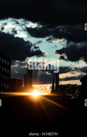 Der Sonnenaufgang der goldenen Stunde zieht sich durch stürmische Wolken, während die Skyline der modernen Stadt in die Silhouette fällt und dramatische Strahlen über den Horizont wirft. Stockfoto