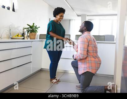 Ein afroamerikanisches Paar, das kniet und lächelt in einer modernen weißen Küche, mit offenem Ringkasten. Romantisch, fest, Zuneigung, Eleganz, Intima Stockfoto