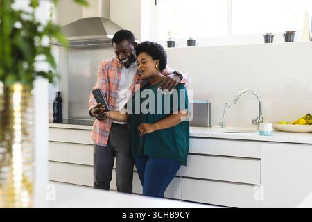 Das afroamerikanische Paar hält ein Smartphone und lehnt sich an die Arbeitsplatte, mit Topfpflanzen und Bananen. Modern, Lifestyle, minimalistisch, innen, aff Stockfoto