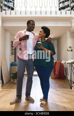 Stehendes afroamerikanisches Paar, das Kaffeetassen im Eingangsbereich hält, mit Umzugskartons. Zuhause, Paar, modern, Innenraum, Lifestyle, gemütlich, DE Stockfoto