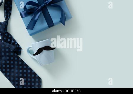 Kreative Grußkarte mit blauer Tasse, Krawatte und Geschenkbox auf hellblauem Hintergrund. Alles Gute Zum Vatertag. Schnurrbart Monat Challenge. Herrenaccessoires Stockfoto