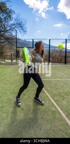Eine junge, starke, glückliche Athletin, lächelnd, kurz davor, einen Padel-Ball zu treffen, ihren Schläger in Position, auf einem Outdoor-Platz mit einer bergigen Landschaft Stockfoto