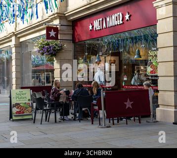 Außenterrasse von Pret a Manger mit Leuten, die an einem sonnigen Tag Speisen und Getränke genießen, neben einer Ladenfront mit hängenden Blumen Bath UK Stockfoto