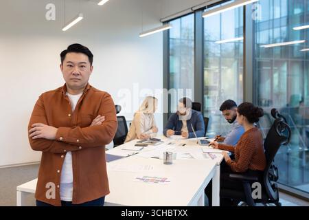 Selbstbewusster asiatischer Manager, der mit überkreuzten Armen ein Geschäftstreffen mit seinem Team in einem hellen, modernen Büro führt und Strategien und pla bespricht Stockfoto